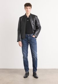 Čierna kožená bunda so stojatým golierom, skombinovaná s tmavomodrými džínami. Model má na sebe čierne tričko s logom "HUGO" a čierne členkové topánky.