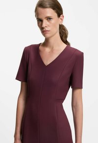 V-kauluspuku syvässä burgundin värisessä kankaassa, jossa on lyhyet hihat ja muodollinen istuvuus. Huomattavat saumurivistykset vartalon kohdalla korostavat siluettia.