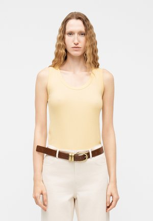 Kobieta o długich, falowanych włosach, ubrana w blady żółty top bez rękawów, białe spodnie oraz brązowy skórzany pasek ze złotą klamrą, stoi na białym tle.