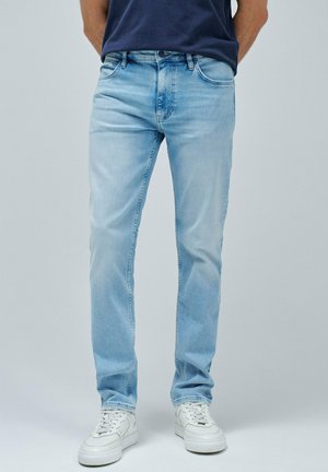 Hombre vistiendo jeans de mezclilla azul claro, zapatillas blancas y una camisa azul marino, de pie con las manos detrás de la espalda sobre un fondo gris liso.