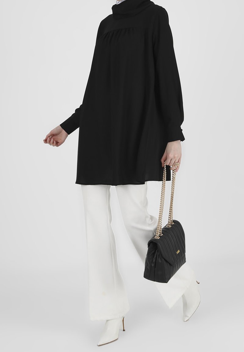 Femme portant une tunique noire à manches longues, un pantalon blanc à jambes larges, des bottes blanches à bout pointu, et tenant un sac à main noir avec des anses en chaîne dorée.