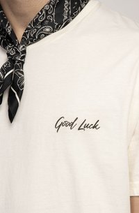 Šviesiai smėlio spalvos medvilninis marškinėlis trumpomis rankovėmis ir išlenktu kaklu, kairėje krūtinės pusėje su užrašu "Good Luck" tamsiomis kursyvomis. Juodas raštuotas bandanas surištas aplink kaklą.
