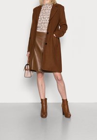 Bruine wollen jas met zwarte knopen over een bloemenblouse, gecombineerd met een bruine leren rok en bruine leren laarzen tot de knie met hakken. Kleine handtas.