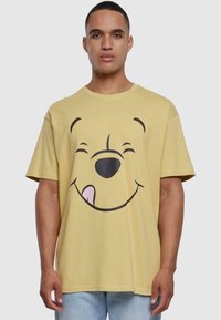 Gul bomull t-skjorte med et stort svart grafisk bilde av en smilende bjørn med rosa tunge og rund halslinning.