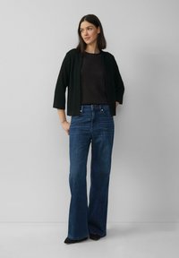 Cardigan en tricot noir à manches trois-quarts, porté sur un haut sombre et un jean large bleu. Des chaussures noires à bout pointu complètent la tenue.
