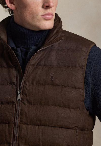 Mężczyzna ubrany w ciemnobrązową pikowaną kamizelkę na granatowym golfie, zwrócony lekko na bok, na neutralnym tle.