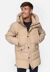 Beige polstret jakke medhette, med glidelås foran, to utvendige lommer og svarte knapper. Glatt tekstur og uformelt design.