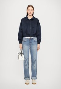 Marineblå bomberjakke med glidelås og elastisk nederkant, kombinert med lyseblå jeans med mønster. Modellen holder en liten hvit håndveske.
