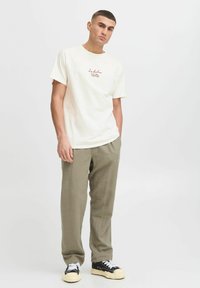 Kremfarget t-skjorte med korte ermer og maroon tekstdesign, kombinert med lyse olivengrønne bukser og svarte joggesko med hvite lisser og beige såler.