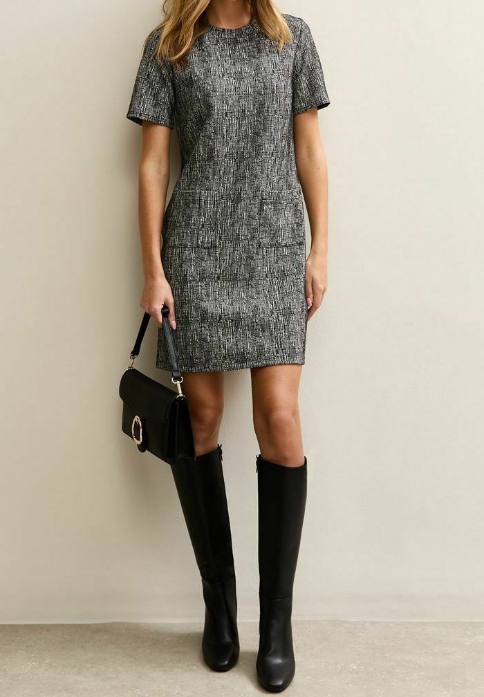 Femme portant une robe grise texturée à manches courtes, des bottes noires montantes et tenant un sac à main noir avec une boucle dorée, debout contre un mur uni.