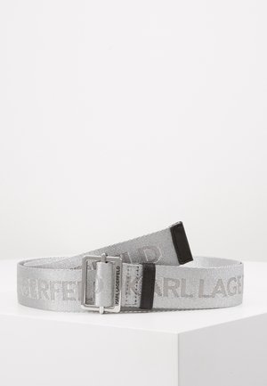 Stříbrný látkový pásek se stříbrnou obdélníkovou sponou, černými koženými detaily a textem "KARL LAGERFELD" tkaným podél délky pásku.