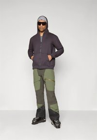 Sweatshirt escura cinza com capuz, fecho de correr frontal e dois bolsos, combinada com calças verde-oliva com remendos e detalhes escuros. Botas de esqui pretas.