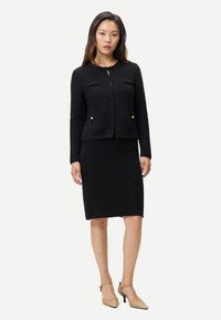 Kvinna som bär en svart texturerad stickad jacka och matchande knälång kjol, kombinerat med beiga lågklackade skor, stående mot en vit bakgrund.