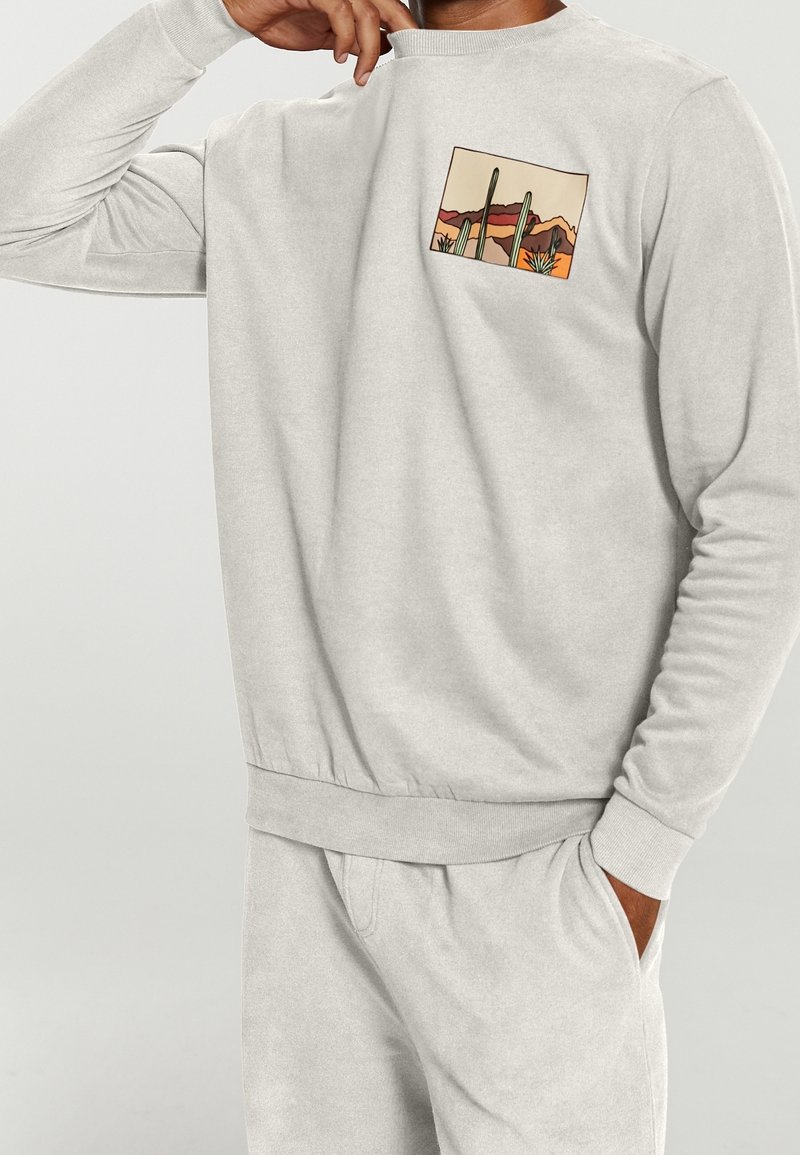 Personne portant un sweat-shirt gris clair avec un petit motif de paysage désertique avec des cactus et des montagnes sur la zone de la poche poitrine.