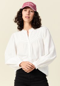 Weißes Hemdbluse mit plissiertem Design und Puffärmeln, kombiniert mit schwarzen Hosen; das Model trägt eine pinke "California"-Kappe.