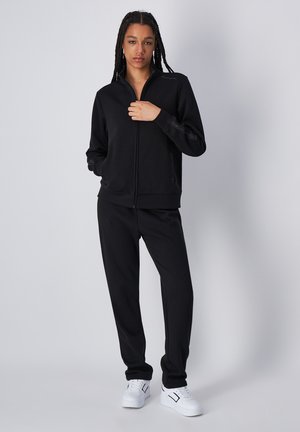 Chaqueta negra con cremallera y cuello alto, puños acanalados y pantalones de chándal negros. Zapatillas blancas completan el conjunto, enfatizando un look monocromático.