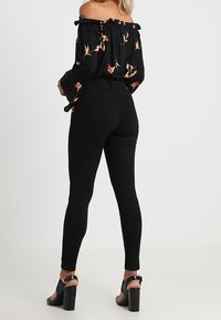 Mujer con blusa floral negra sin hombros, pantalones ajustados negros de cintura alta y sandalias de tacón negras, de pie de espaldas.