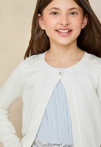 Cardigan en tricot blanc avec des accents froncés sur les épaules et un petit fermoir au niveau du col, superposé sur une robe plissée bleu clair.