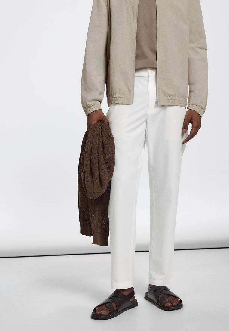 Personne portant une veste beige, une chemise taupe, un pantalon blanc et des sandales foncées, tenant dans une main un pull marron plié.