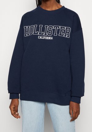 Mørkeblå crewneck-sweatshirt med teksten "HOLLISTER CALIFORNIA", båret med lyseblå jeans på en person med mørk hud og flettet hår.