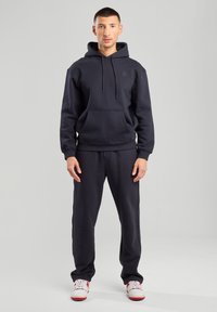 Felpa blu scuro con tasca frontale, cordoncino regolabile e polsini a coste, abbinata a pantaloni coordinati e scarpe da ginnastica bianche.