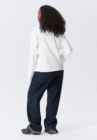 Bílá košile s dlouhým rukávem v kombinaci s tmavě modrými volnými džíny a černými teniskami. Viditelné prošití na kalhotách přidává texturu.