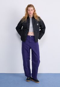 Schwarze Jacke mit Druckknopfverschluss, zwei Brusttaschen und einem Logo-Patch, kombiniert mit lila weiten Hosen und schwarzen Schuhen mit Gummisohlen.