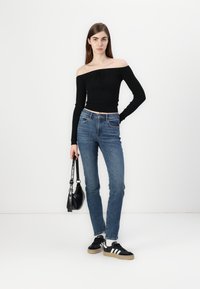 Black off-the-shoulder ribbed sweater with long sleeves, paired with blue straight-leg jeans and black sneakers with white stripes. Black handbag.