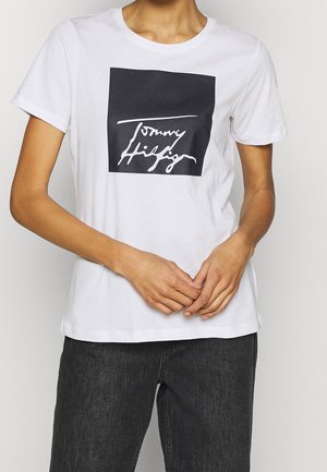 Persoon draagt een wit T-shirt met korte mouwen en een zwart vierkant met wit cursief logo op de borst, gecombineerd met donkergrijze spijkerbroek.