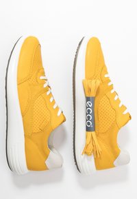 Paire de baskets en cuir jaune avec lacets et semelles blanches, une chaussure présentée avec des lacets jaunes supplémentaires maintenus par une bande de marque.