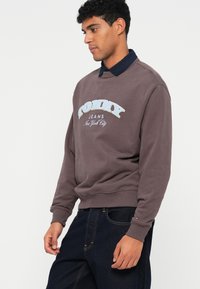 Fialová mikina s okrúhlym výstrihom, s bielym a modrým logom "TOMMY JEANS New York City" v zakrivenom dizajne. Viditeľné tmavé džínsy.