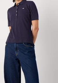 Personne portant un polo à manches courtes bleu marine avec des boutons blancs et un logo brodé, assorti à un jean denim bleu foncé.
