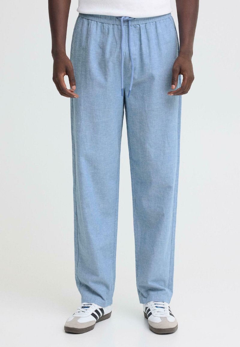 Homme portant un pantalon léger bleu ciel à cordon de serrage et des baskets blanches à rayures noires, debout devant un fond uni.