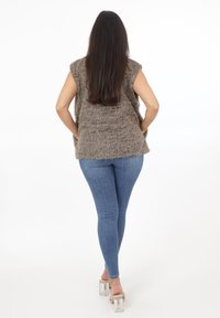 Faux fur väst i brunt, kortärmad, lager av textur. Stylistisk med blå jeans och klara blockklacksandaler. Vit bakgrund.