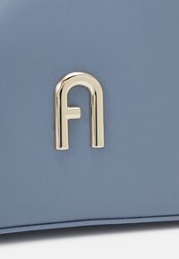 Sininen nahkainen laukku, jossa on sileä pinta ja omaavassa korostetun kultaisen metallilogon yksityiskohta. Muotoilu on tyylikäs ja moderni.