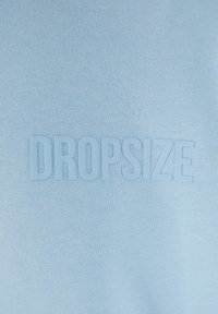 Vaaleansininen huppari, jossa on sileä pinta, etuosassa on kohokuvioitu teksti "DROPSIZE". Yksinkertainen muotoilu.