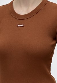 Κοντινό ενός ατόμου που φοράει καφέ ριπ T-shirt με μια μικρή ορθογώνια ετικέτα "HUGO" στο κέντρο κάτω από το λαιμόκοψη.