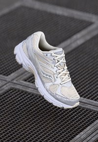 Chaussure de course légère en crème et gris, avec une tige en maille, une semelle épaisse et un système de laçage distinctif avec un motif texturé.