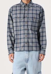 Camisa de franela a cuadros azul y gris con cuello de botón, dos bolsillos en el pecho y botones marrones, combinada con pantalones de mezclilla azul claro.