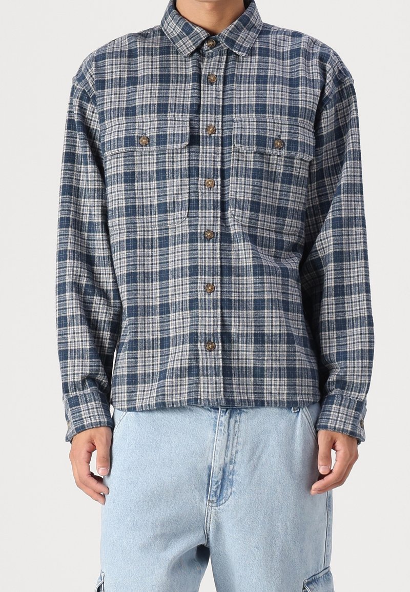 Camisa de franela a cuadros azul y gris con cuello de botón, dos bolsillos en el pecho y botones marrones, combinada con pantalones de mezclilla azul claro.