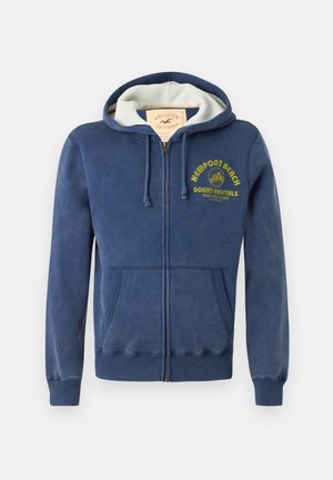 Mornarsko modra zip-up kapuca, izdelana iz mešanice bombaža, z obloženim robom, dvema sprednjima žepoma in rumenim potiskanim besedilom z dizajnom palme.