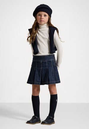 Jupon en denim plissé, gilet en tricot bleu marine, col roulé blanc, chaussettes montantes et chaussures noires. Les accessoires incluent un béret bleu marine.