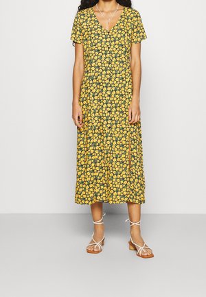 Kvinde iført en gul blomsterprintet midi-kjole med korte ærmer og en sideslids, stylet med hvide sandaler med lav hæl og stropper.