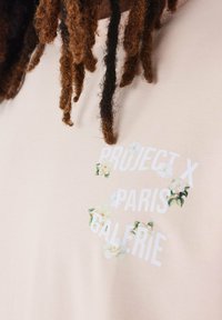 Lichtroze t-shirt met een bloemmotief en de tekst "PROJECT X PARIS GALERIE" in het wit. Zachte stof, korte mouwen en een ontspannen pasvorm.