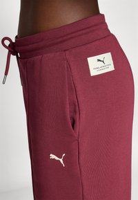 Burgundia puuvillased joggers, millel on nööriga vöökoht, küljetaskud ja valge Puma logo plekk vasakul reiel. Sile tekstuur.