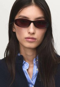 Gafas de sol con lentes oscuros en forma ovalada y finos marcos negros. Llevadas con una camisa de cuello azul debajo de un cárdigan azul marino. Textura suave, diseño ligero.