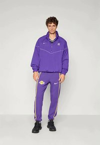 Lilla Nike treningsdrakt med jakke med kvartglidelås og matchende bukser med Lakers-logo og gule sidestriper. Svarte joggesko er brukt.