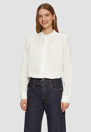 Blouse blanche à manches longues avec un col froncé et un devant boutonné, associée à un pantalon en denim foncé taille haute avec une ceinture.