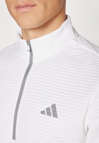 Pullover bianco con colletto alto, caratterizzato da una zip frontale a metà. Tessuto con texture a costine; include il logo grigio di Adidas sul petto.