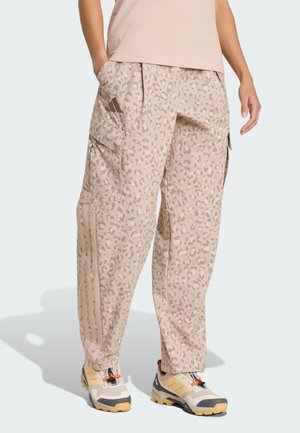 Person iført beige mønstrede cargobukser med sidelommer og beige vandresko med orange snørebånd på lys baggrund.